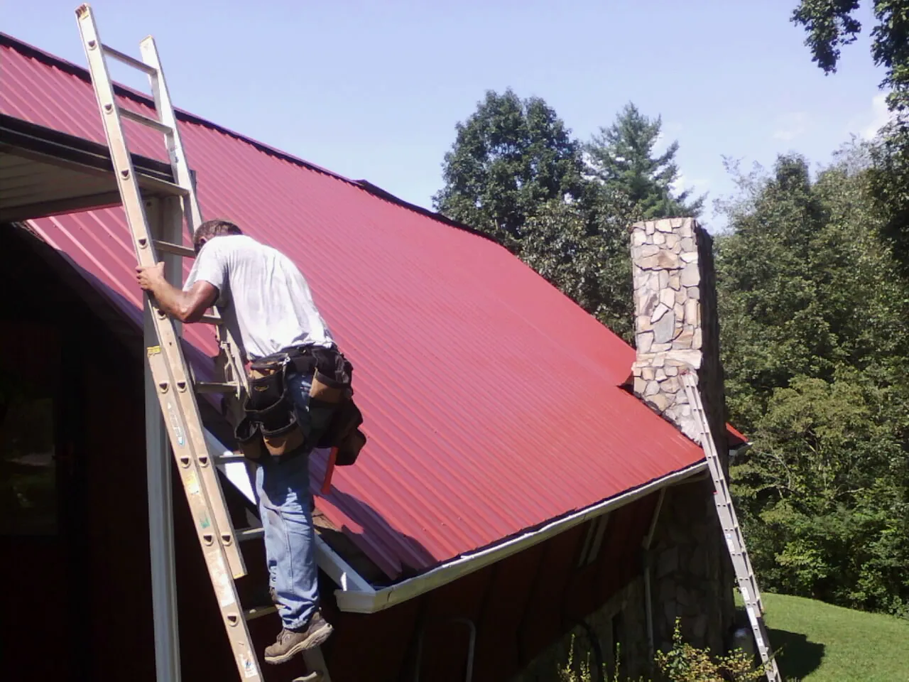 Roofer on a ladder beside a red metal roof and stone chimney — Soffit & Fascia Repair by Roofing Brothers in Houlton